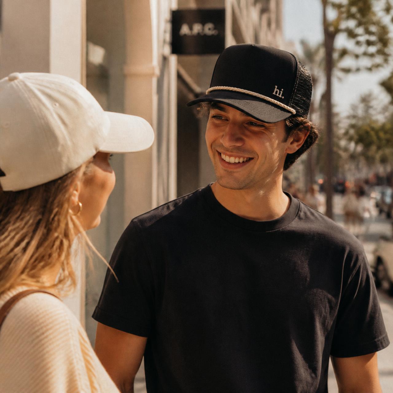 A young couple smiling at each other on a sunny sidewalk — he's wearing the uSayHi black rope trucker hat with a small cream 'hi.' embroidered on the front